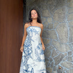 A woman wearing a strapless white maxi dress with a blue nautical print of starfish, palm trees, and sailboats, standing in front of a stone wall and wooden door.