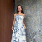 A woman wearing a strapless white maxi dress with a blue nautical print of starfish, palm trees, and sailboats, standing in front of a stone wall and wooden door.
