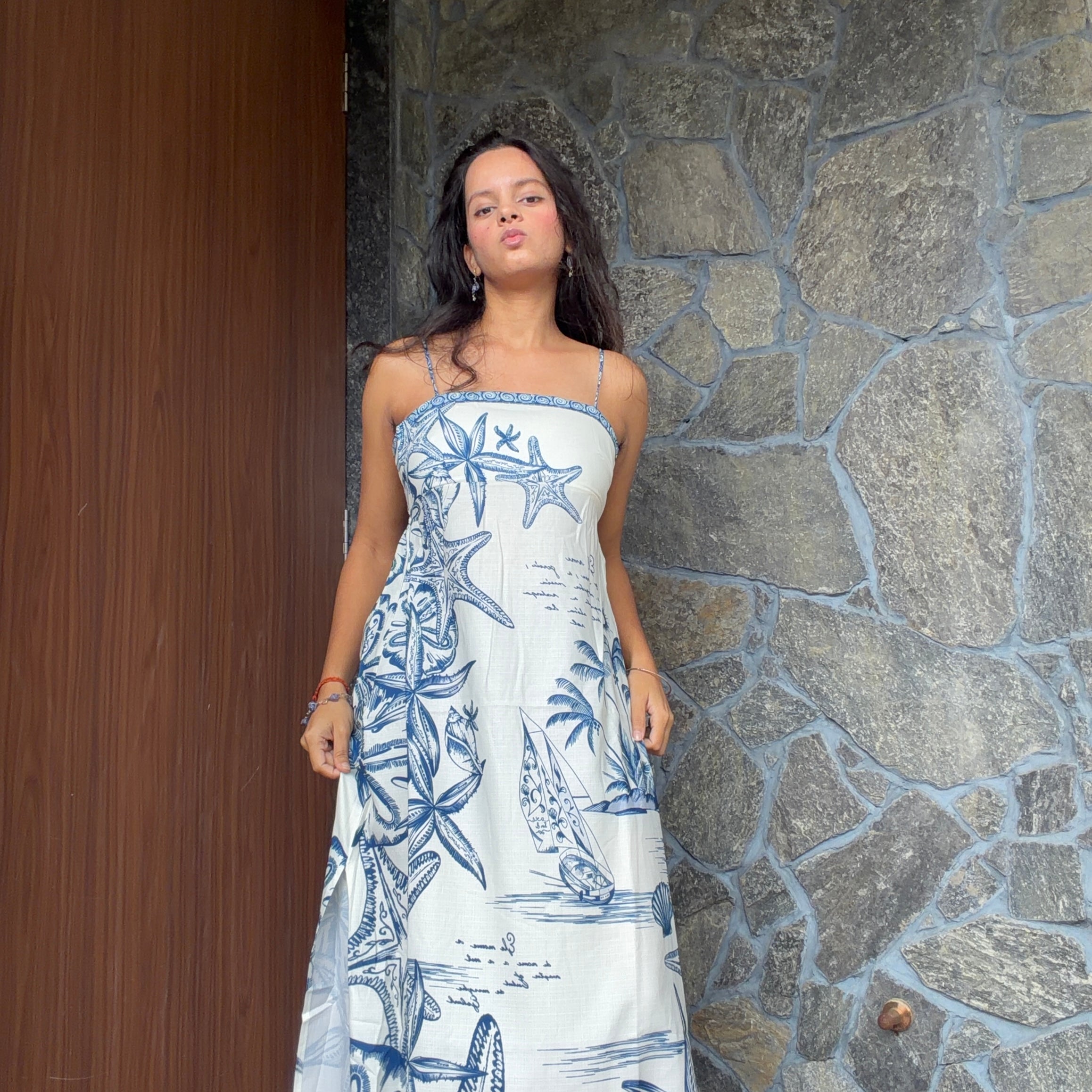 A woman wearing a strapless white maxi dress with a blue nautical print of starfish, palm trees, and sailboats, standing in front of a stone wall and wooden door.