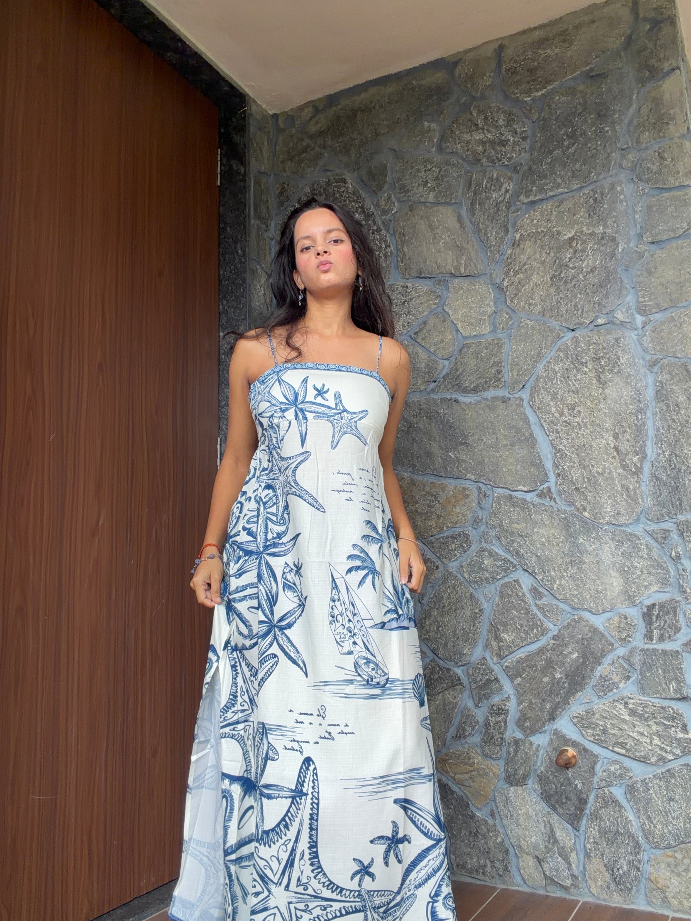 A woman wearing a strapless white maxi dress with a blue nautical print of starfish, palm trees, and sailboats, standing in front of a stone wall and wooden door.